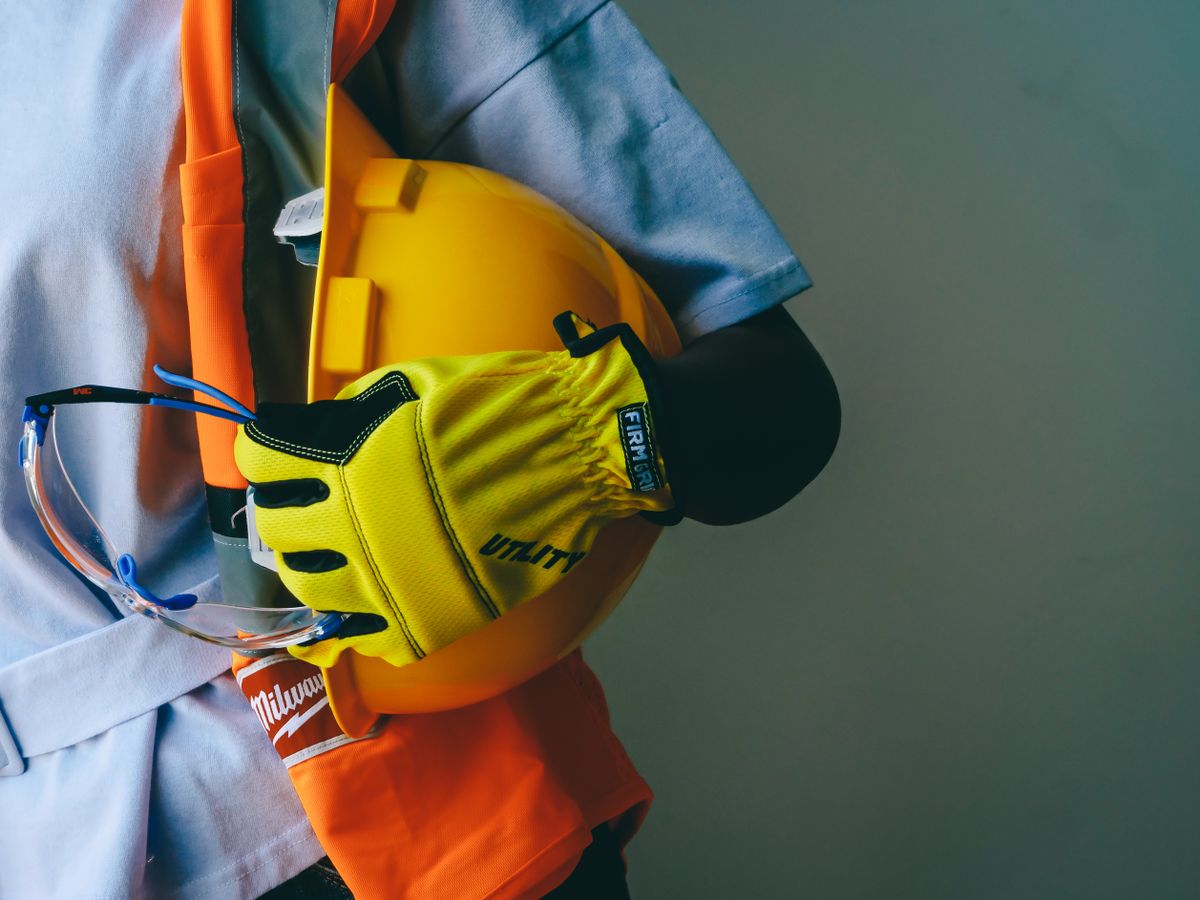 A worker in a hi-vis vest holding a hard hat, gloves and goggles