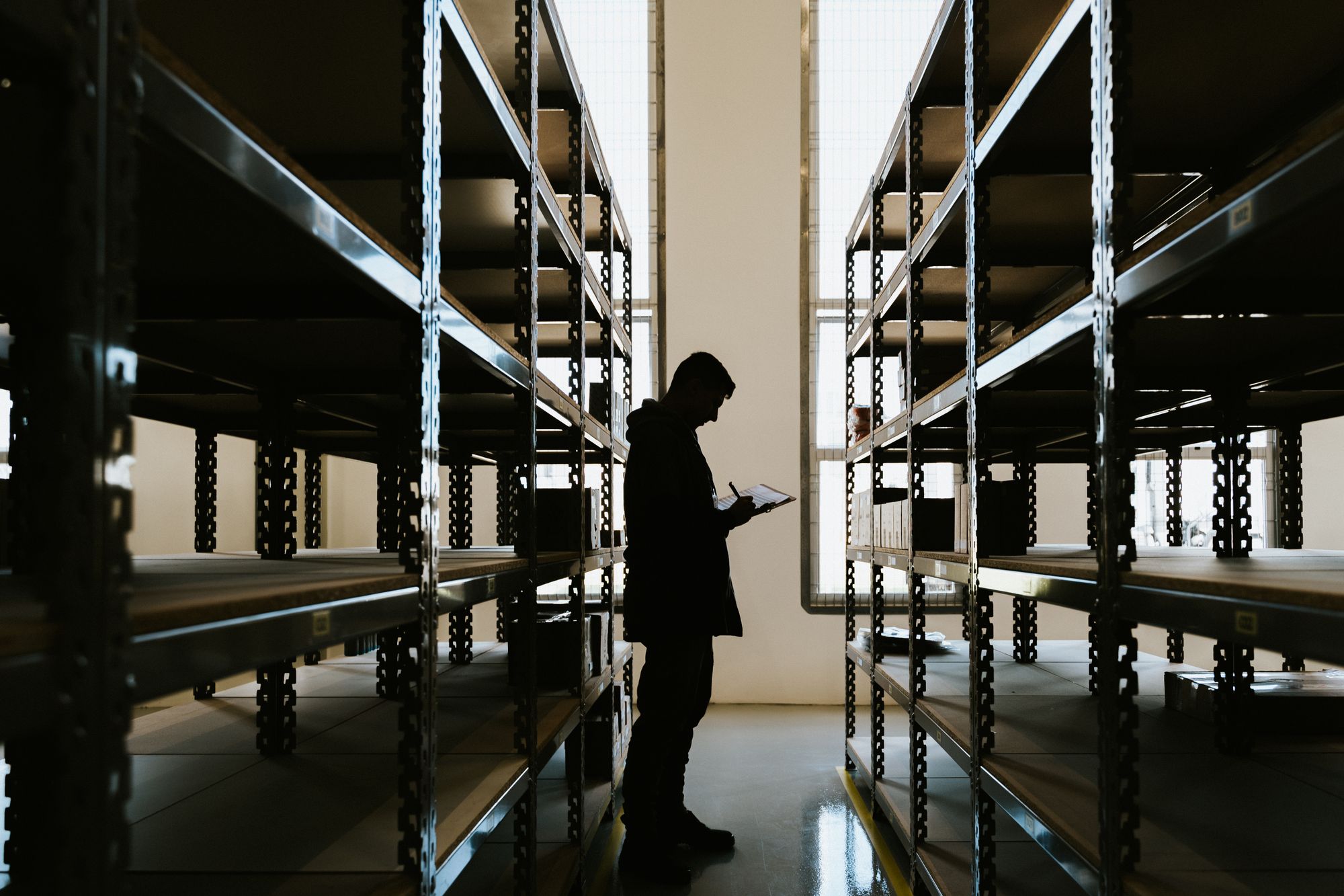A stock controller checking inventory against a clipboard between warehouse racking